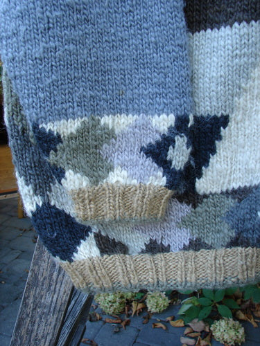 Tara Handknits Diamond Button Oversized Cardigan Geometric Naturals OSFA draped on a bench, showcasing its intricate blocky diamond and striped pattern, ribbed hem, and flap pocket, capturing its vintage charm.