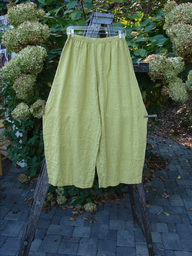 Barclay NWT Linen Alchemist Pant Unpainted Citron Size 0 hanging on a wooden frame, showcasing wide legs, sectional panels, and dual front pockets against a lush, garden backdrop with stone paving.
