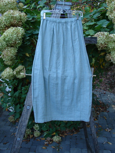 2000 Cross Dye Linen Contour Skirt Unpainted Meadow Size 0 hanging on a wooden frame, showcasing its full elastic waistband, vertically paneled sides, and darted reverse bell bottom accents in a garden setting.
