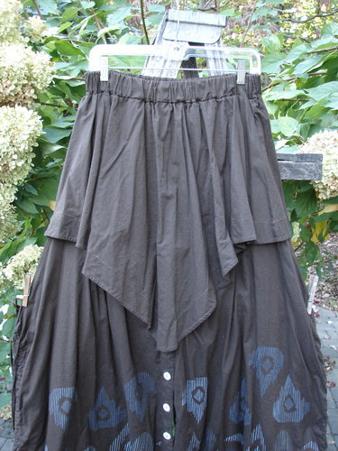 Barclay Batiste Split Button Pant Skirt Celtic Mocha Size 0, featuring a dark-brown tiered design with an elastic waistband, draped overlay, and buttoned blue-patterned hem, hanging on a clear plastic hanger outdoors.