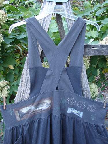 1995 Overall Jumper Fish Space Briquette OSFA: A sleeveless dress with crisscross straps, gathered skirt, and decorative waistband with fish and spiral motifs, photographed against a wooden ladder amidst green foliage.