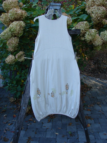 2000 Cotton Hemp Libby Jumper Falling Leaf Dove Altered Size 1: White sleeveless scoop-neck jumper with bubble hem and embroidered flowers, displayed on a wooden A-frame with hydrangeas in the background, highlighting its elegant drape.