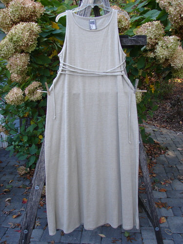 1998 Linen Knit Wrap Dress Unpainted Natural Size 1 hangs elegantly on a wooden A-frame, showcasing its slender drape, thin waist cords, and graceful rounded neckline against a natural garden backdrop.