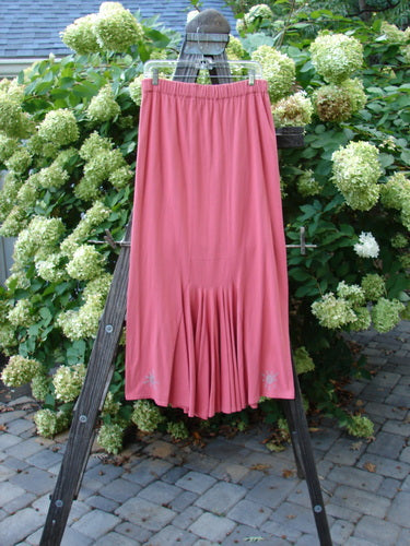 A 1995 Kick Pleat Skirt in Papaya, part of the Winter Resort Collection, is shown. The skirt features a full elastic waist, elongating shape, rear kick pleat, and painted accents on the waistband. The image shows a pink skirt on a wooden ladder, with a close-up of a plant and a wooden post.