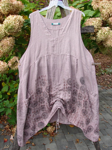 Barclay Linen Pleated Pocket Pinafore Mandala Soft Mauve Size 2 features a scooped neckline, side pockets, and gathered hem with drawstring. Displayed outdoors, showcasing subtle floral motifs on medium-weight linen.