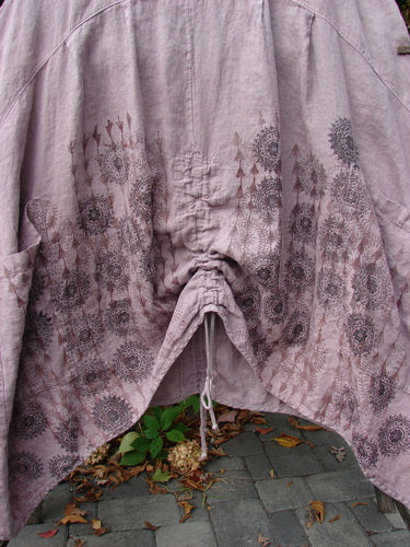 Barclay Linen Pleated Pocket Pinafore in Soft Mauve features a gathered draw-string detail, mandala floral print, and dual vertical drawcords, showcased in an outdoor setting over cobblestones.