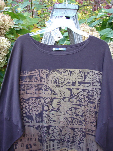 Barclay NWT Modern Square Top Metallic Village Loam Ogo OSFA displayed outdoors on a ladder, showcasing a tan/gold village-themed screen-print on its dark organic cotton fabric with dolman shoulders and rounded neckline.
