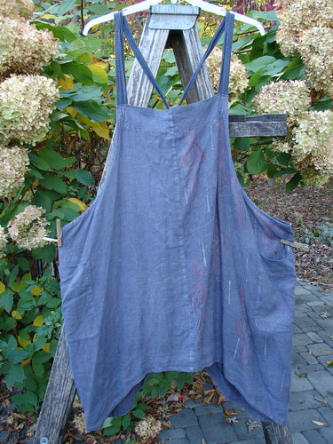 Barclay Linen Apron Jumper Celtic Rain Raven Size 2 hanging on a wooden ladder, features a sleeveless design, asymmetrical hem, and unique striped shoulder straps with side pockets.