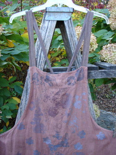 Barclay Linen Apron Jumper Rose Rusty Bucket Size 2, featuring a faded floral pattern, adjustable striped shoulder straps, banded upper bib, and two wrap drop side pockets, hangs on a weathered step-ladder.