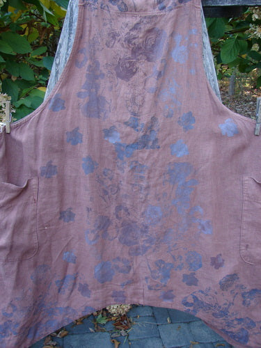 Barclay Linen Apron Jumper Rose Rusty Bucket Size 2, featuring two large pockets, adjustable striped straps, and a unique floral pattern, hangs outdoors, showcasing its flared shape and medium-weight linen fabric.
