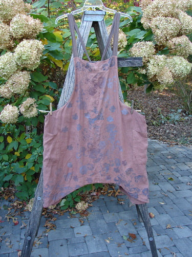 Barclay Linen Apron Jumper Rose Rusty Bucket Size 2 displayed outdoors on a wooden ladder, features a sleeveless design, adjustable striped straps, two wrap drop side pockets, and a varying hemline.