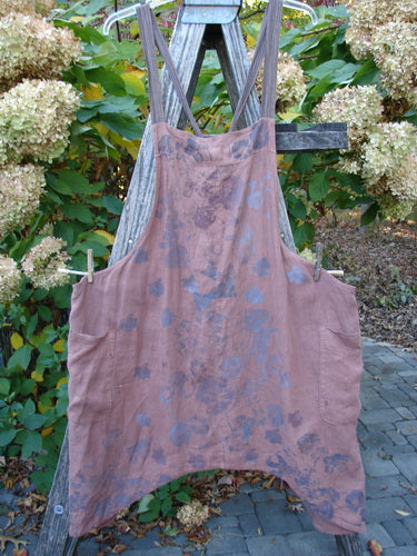 Barclay Linen Apron Jumper Rose Rusty Bucket Size 2 draped over a wooden stepladder outdoors, featuring wide side pockets and crossed adjustable shoulder straps with a mottled rose and gray/blue pattern.