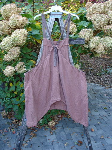 Barclay Linen Apron Jumper Rose Rusty Bucket Size 2: A sleeveless, dusty pink apron dress with crisscross tie straps and two side pockets, hanging outdoors on a wooden stand.