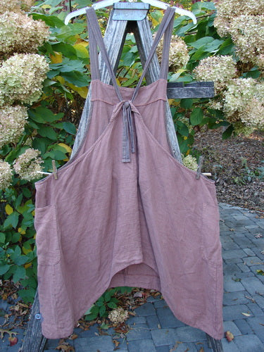 Barclay Linen Apron Jumper Rose Rusty Bucket Size 2, featuring a medium-weight linen blend with wide cross-back straps, deep side pockets, and a banded bib, displayed on a rustic ladder outdoors.