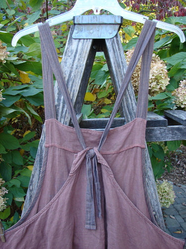 Barclay Linen Apron Jumper Rose Rusty Bucket Size 2 displayed on a wooden trellis, featuring uniquely striped adjustable halter straps, a gathered bodice, front pocket, and flowing hemline, perfect for expressing creative individuality.