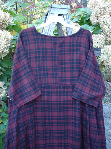 Barclay Flannel Bottom Bell Pocket Dress Merlo Plaid Size 2, displayed on a white hanger over a wooden trellis, showcasing its loose fit, rounded neckline, and wide three-quarter sleeves.