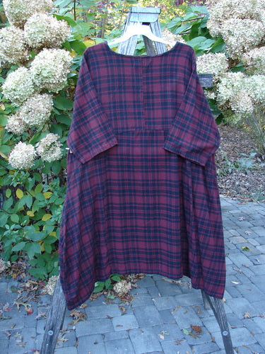 Barclay Flannel Bottom Bell Pocket Dress in Merlo Plaid Size 2 is displayed on a wooden easel, showcasing its loose fit, rounded neckline, and three-quarter sleeves, set against a garden backdrop.