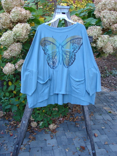 Barclay NWT Two Pocket Mariposa Pullover Butterfly Sea OSFA displayed on a wooden step-ladder, featuring a large butterfly graphic, wide three-quarter sleeves, and two patch pockets in a garden setting.