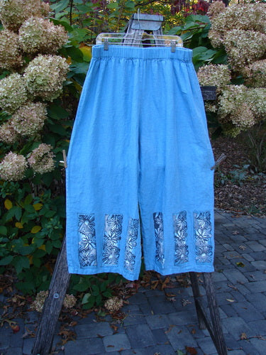 Barclay Linen Wide Leg Crop Pant Daisy Trio Cornflower Size 2, featuring loose-fitting, cropped design with elastic waistband and white floral prints on hem, displayed outdoors on a wooden rack amidst garden foliage.