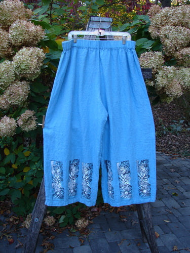Barclay Linen Wide Leg Crop Pant Daisy Trio Cornflower Size 2, featuring loose-fitting, calf-length design, elastic waistband, and decorative black-and-white floral panels along the hem, hanging outdoors.