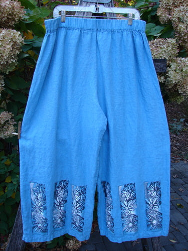 Barclay Linen Wide Leg Crop Pant Daisy Trio Cornflower Size 2, featuring wide cropped legs, floral-print hem panels, and an elastic waistband, displayed hanging outdoors against lush foliage.