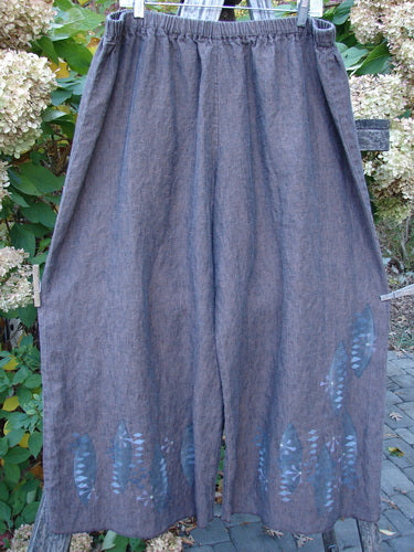 Barclay Cross Dye Linen Wide Leg Crop Pant Floral Sphere Wood Size 2, showcasing wide-legged, gray-brown pants with an elastic waistband and floral sphere design near the hem, hangs outdoors against a garden backdrop.