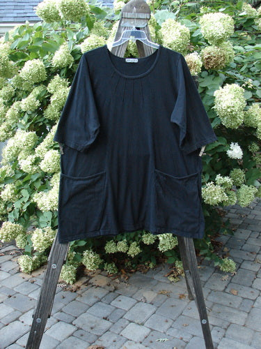 Barclay Sunrise Pin Tuck Pocket Tunic Unpainted Black Size 2 displayed on a wooden rack, highlighting its A-line shape, deep rounded neckline, short sleeves, and unique front pockets.