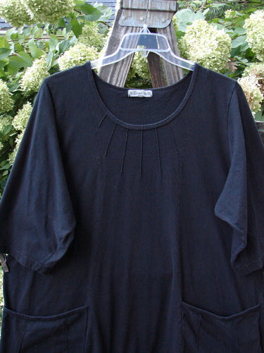 Barclay Sunrise Pin Tuck Pocket Tunic Unpainted Black Size 2 displayed on a clear plastic hanger, highlighting its deeper rounded neckline, A-line shape, short sleeves, and pin-tucked front pockets.