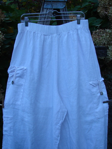 Barclay Linen Button Tab Tool Pocket Pant Unpainted Orchid Size 2: Loose-fitting white pants with elastic waistband, flap-pocket detailing, and wide billowy legs, displayed on a hanger amidst outdoor foliage.