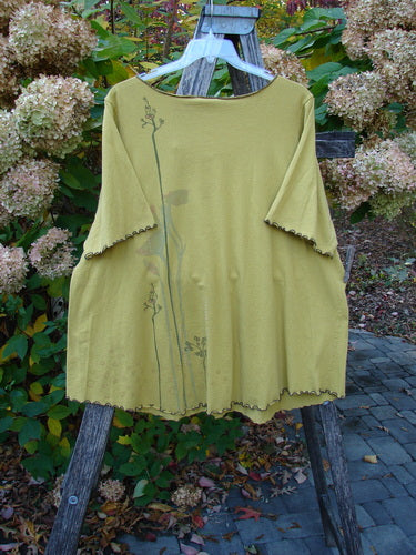 Barclay Gather Twinkle Top Berry Rise Yellow Olive Size 2: A loose-fitting, three-quarter-sleeve tunic with floral stems and scalloped edges, displayed outdoors on a wooden stepladder among hydrangeas.