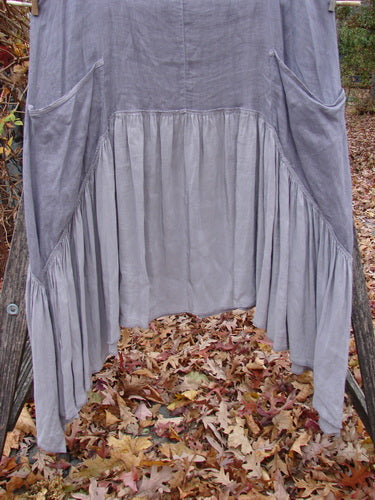Barclay Gauze Silk Summer Frock Jumper Unpainted Grey Haze Size 1 draped over a wooden rail, featuring a scooped neckline, exterior wrap pockets, and a ruffled silk lower skirt in a serene outdoor setting.