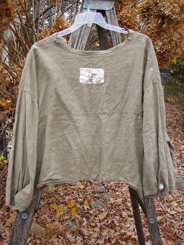 Magnolia Pearl Linen Crop Artist's Pocket Pullover Light Army OSFA displayed on a hanger over a ladder, featuring billowy three-quarter sleeves, dual pockets, and a unique fabric patch below the neckline.