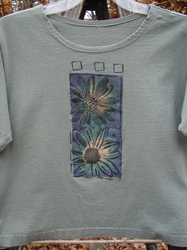 1996 Crop Tee Floral Butterfly Cricket Size 1: A boxy, sage-green T-shirt with short sleeves, featuring vertically printed daisies and butterflies, displayed outdoors. Vintage Blue Fish Clothing, organic cotton, spring collection.
