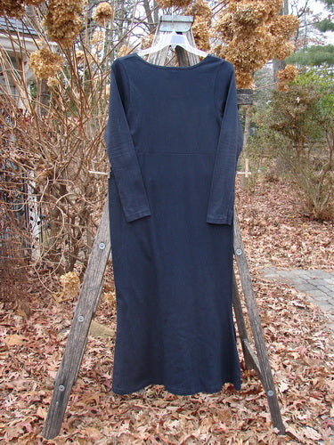 1992 Pedestal Dress Unpainted Black Size 1, a vintage long-sleeved, ankle-length dress with a unique tapered shape and deeper rounded neckline, elegantly displayed on a hanger against a garden backdrop.