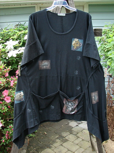 Barclay Double Pocket Bounce Tunic Dress Cat & Mouse Black Size 2 featuring cartoon characters, wrap-around side seams, oversized front pockets, scooped hemline, and oval neckline, displayed on a stone floor backdrop.