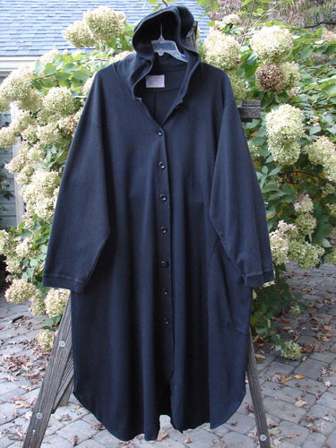 Barclay Interlock Hooded Dahlia Coat Unpainted Black OSFA hanging on a wooden rack, showcasing its large hood, oversized pocket, and distinctive A-line shape with ribbed cuffs.