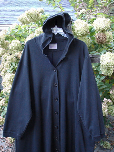 Barclay Interlock Hooded Dahlia Coat Unpainted Black OSFA displayed on a hanger, showcasing its A-line shape, large hood, ribbed cuffs, and oversized pocket with decorative buttons.