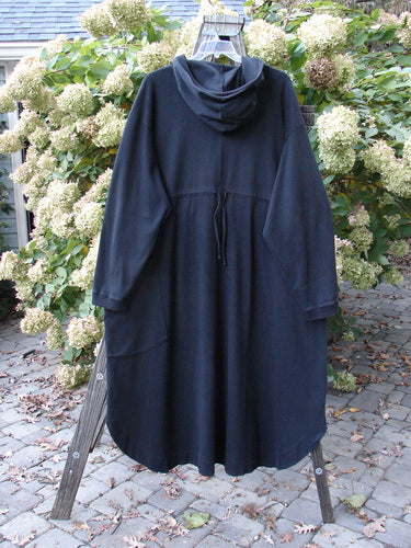 Barclay Interlock Hooded Dahlia Coat Unpainted Black OSFA on a wooden rack, featuring a large cozy hood, ribbed cuffs, oversized pocket, and unique A-line shape with rounded side vents.