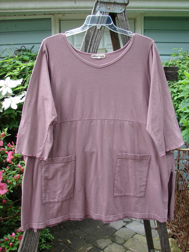 Barclay Empire Vented Pocket Top in Dusty Plum on hanger, featuring a rounded neckline, tall vented sides, two exterior drop pockets, and three-quarter sleeves, showcasing its A-line shape and downward curved empire waist seam.