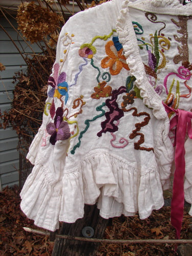Magnolia Pearl Embroidered Linen Ruffle Ribbon Bolero White OSFA, featuring vibrant floral embroidery, ruffled hem and sleeves, and a pink ribbon closure, displayed outdoors against dried leaves and branches.