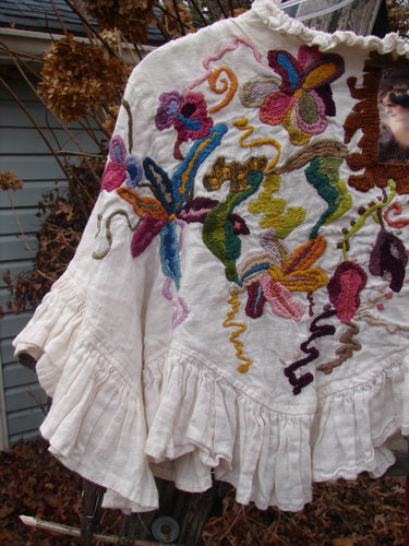 Magnolia Pearl Embroidered Linen Ruffle Ribbon Bolero White OSFA drapes on a hanger, showcasing intricate floral embroidery, a ruffled hem, and a silken ribbon closure, embodying artistic elegance.