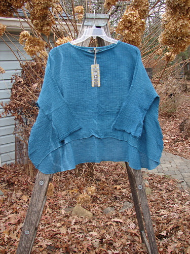 Barclay NWT Crinkle Gauze Top Mottled Teal OSFA displayed on a clear hanger, showcasing its boxy cut, three-quarter dolman sleeves, and high-low hem, set against a rustic outdoor backdrop.