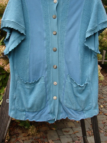 Barclay Celtic Moss Hooded Pocket Cardigan in Dusty Aqua, displayed on a rack, showcasing its short sleeves, floppy hood, button front, and two lower exterior pockets.