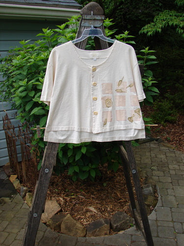 2001 Button Crop Top Game Garden Natural Size 1 displayed on a wooden ladder under a tree, showcasing its flowing boxy style, varying hemline, and unique game-themed paint pattern.