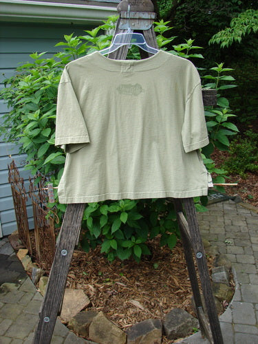 2001 Button Crop Top Junk Drawer Kelp Size 0 displayed on a wooden easel; features wide short sleeves, boxy style, varied hemline, and unique buttons.