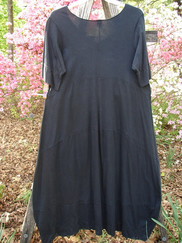 Barclay Gather Sectional Banded Petal Dress Unpainted Black Size 1, displayed on a wooden ladder, features a round neckline, elbow-length sleeves, and a unique dippy hemline against flowering shrubs.