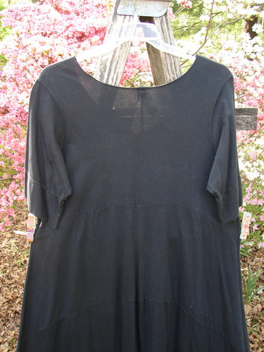 Barclay Gather Sectional Banded Petal Dress Unpainted Black Size 1 displayed on a wooden hanger, featuring elbow-length sleeves and a scooped neckline, with a gentle garden backdrop.