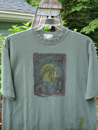 2000 Short Sleeved Tee Celtic Letter Park Size 1 displayed on a hanger, featuring drop shoulders, a thicker ribbed neckline, and a longer straight shape with classic Celtic letter theme paint.