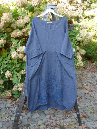 Barclay Linen Rear Tab Pocket Dress in Dusty Navy on a wooden rack, showcasing its wider neckline, pleated sleeves, and dusk floral theme with angular flop pockets.