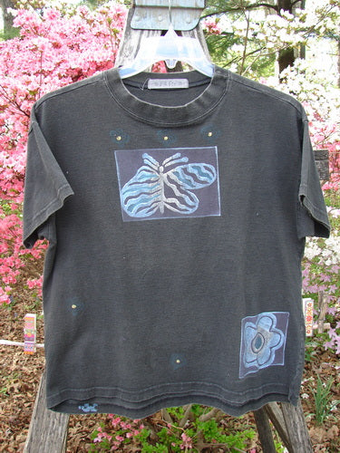 1997 Short Sleeved Tee Butterfly Ebony Size 0 hangs on a fence, featuring a zebra-striped butterfly and floral motifs in blue and gray. It's part of Bluefishfinder.com's vintage collection, showcasing unique artistic designs.